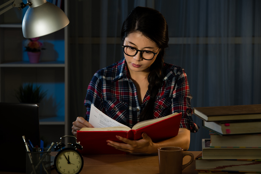 Young student at home desk reading studying at night with pile of books and coffee cup preparing exam in university education concept in dim light. mixed race asian chinese model.