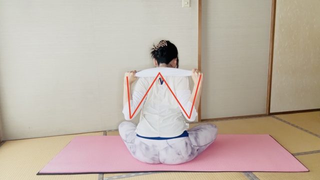 座った姿勢でタオルを肩の後ろに引き、肩甲骨を寄せるエクササイズをする女性